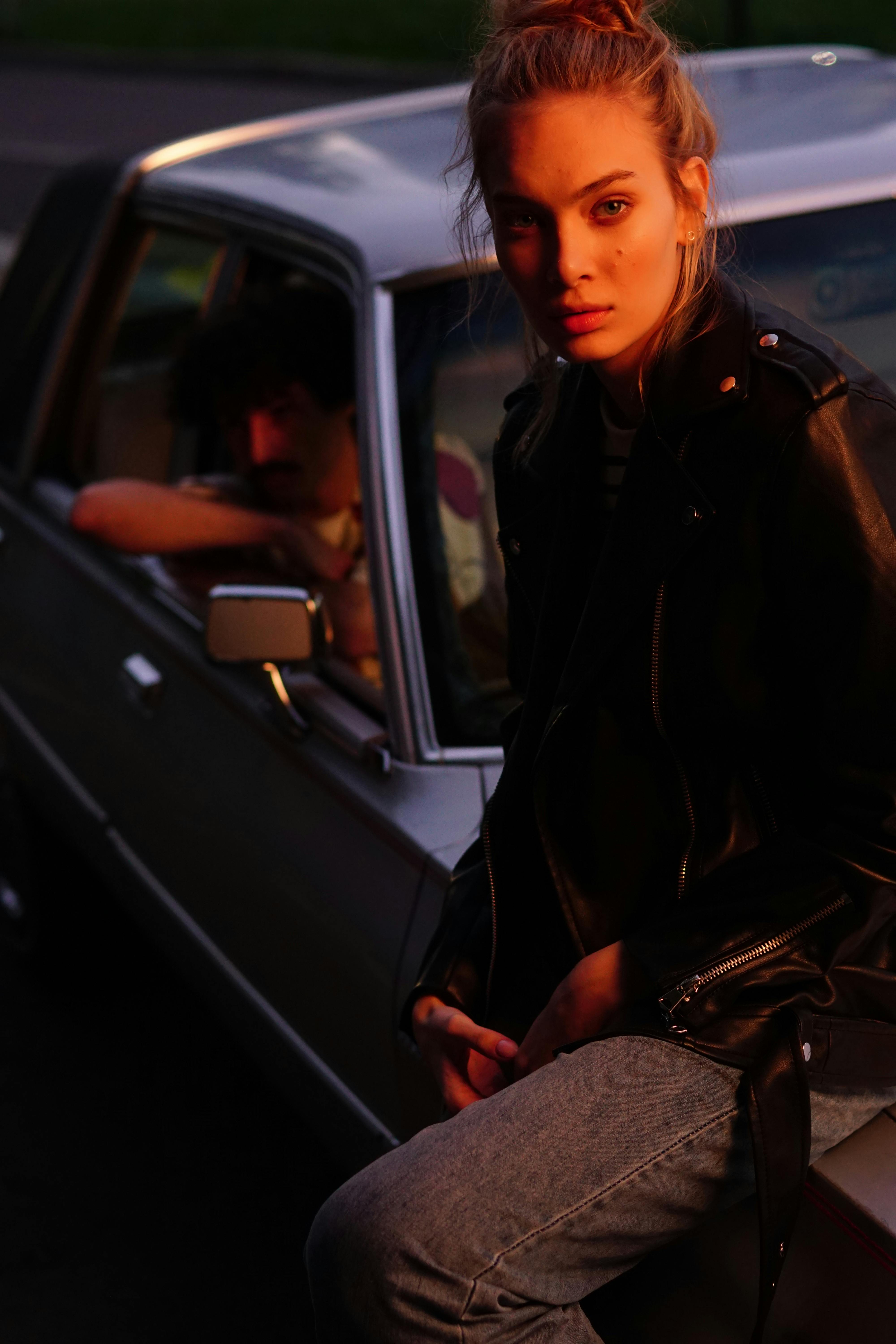 woman in black leather jacket and denim jeans sitting on a car hood