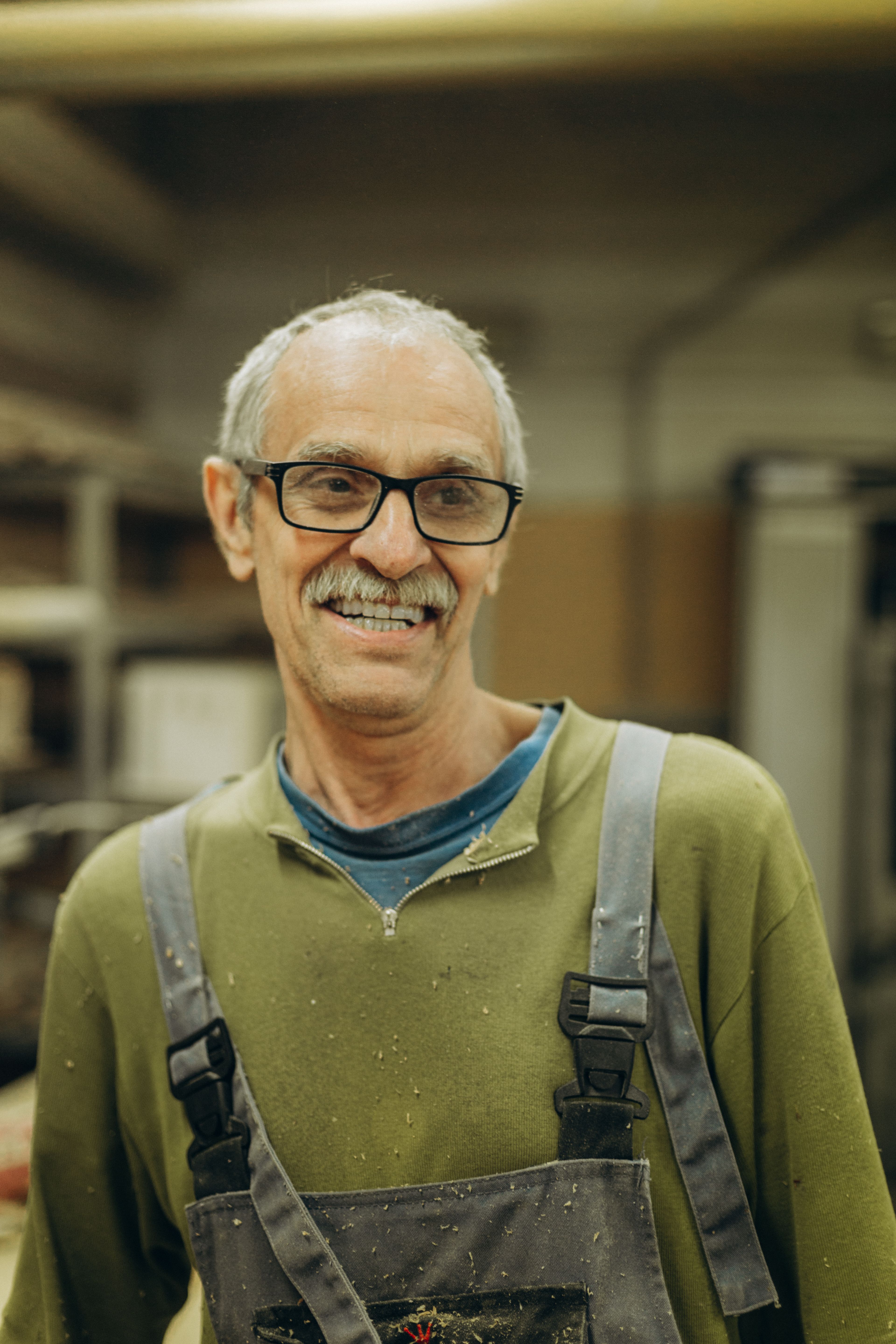 Senior man wearing eyeglasses and overalls smiling indoors in a workshop.