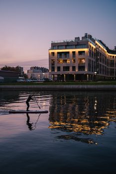 A woman paddleboards on calm water reflecting modern architecture at sunset.