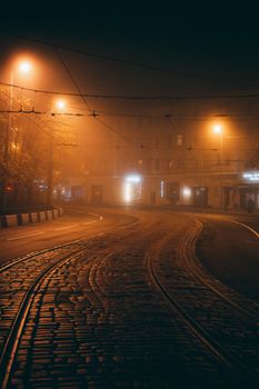 A foggy, illuminated urban street at night with cobblestones and tram tracks, creating a dramatic and moody atmosphere.