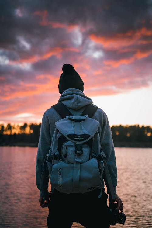 Person in Gray Hoodie Standing Near Body of Water during Sunset