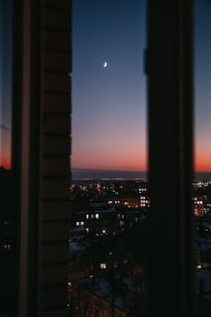 A beautiful cityscape at dusk framed by a window, featuring a crescent moon and vibrant evening sky.