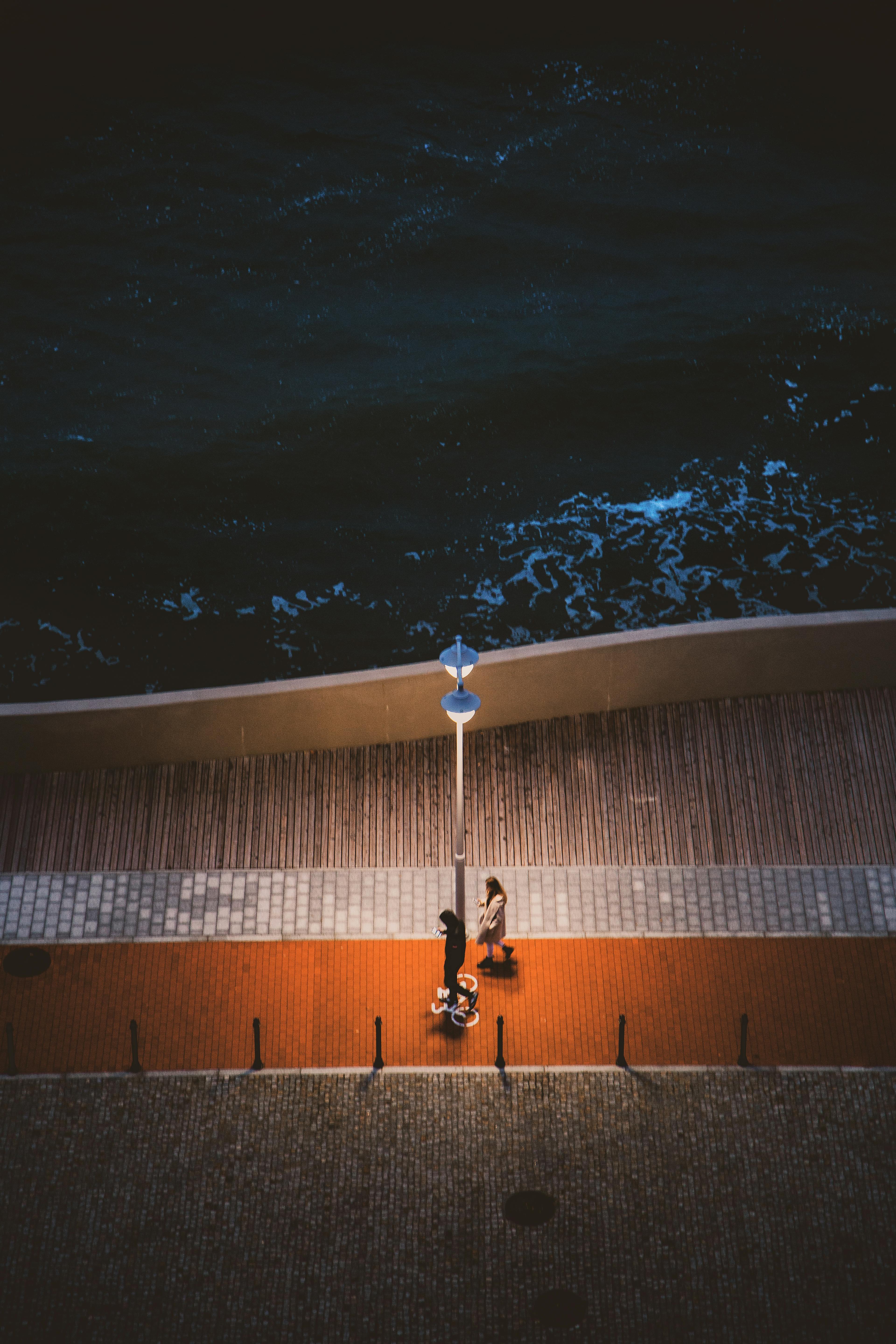 A couple walking on a brightly lit pier by the ocean at night, viewed from above.