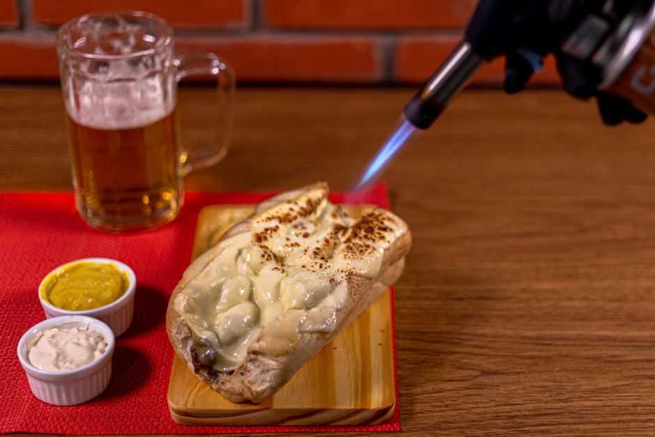 Close-up of a cheesy sandwich being grilled with a torch, served with beer and sauces.