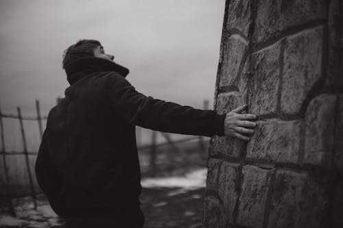Black and White Photography of Man Standing and Touching the Wall of a Building