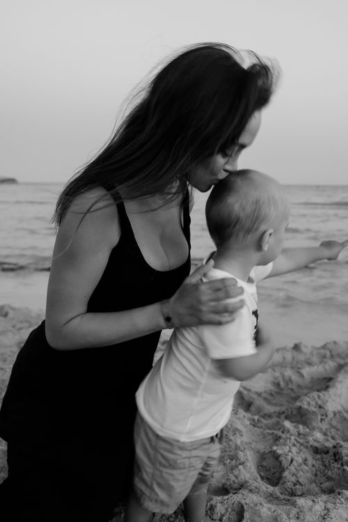 Free A mother lovingly kisses her child on the seashore in a black and white setting, evoking warmth and connection. Stock Photo