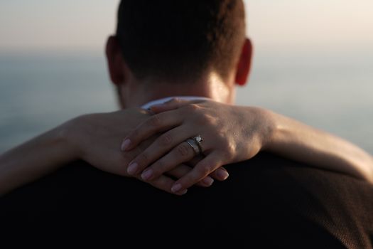 Close-up of a couple embracing with hands featuring diamond and wedding rings, against a serene seascape.