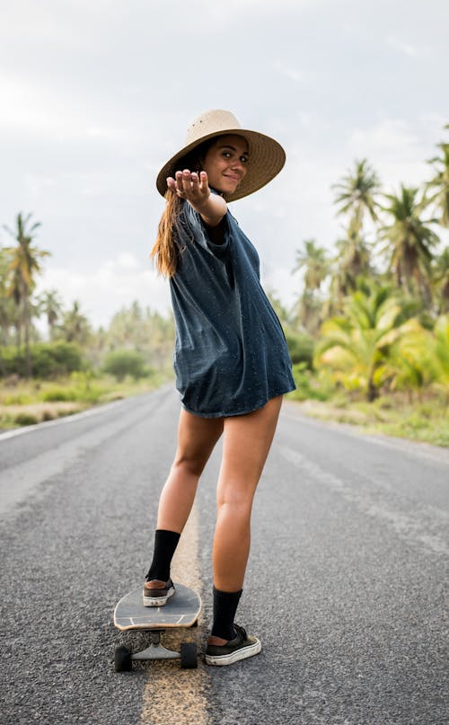 Woman in Blue Long Sleeve Shirt and Black Skirt Wearing Brown Sun Hat Standing on Road