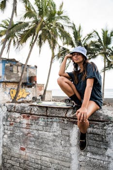 A young woman in casual wear sits on a wall with her longboard near a tropical beach.