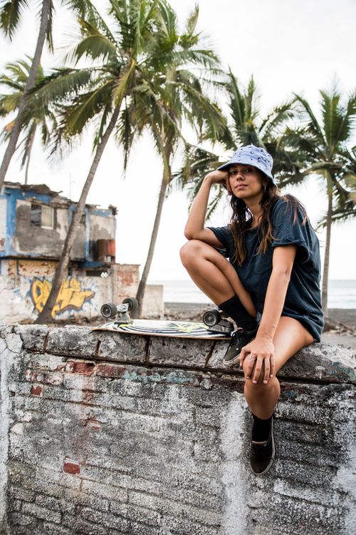 Free A young woman in casual wear sits on a wall with her longboard near a tropical beach. Stock Photo
