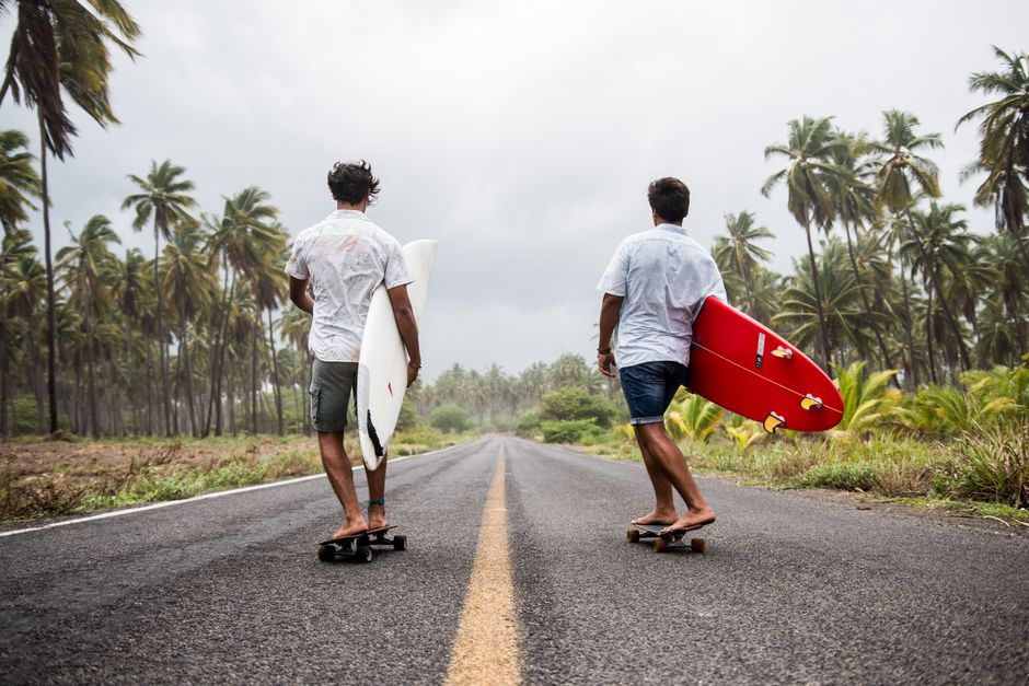 Two young men skateboarding with surfboards on a tropical road lined with palm trees.