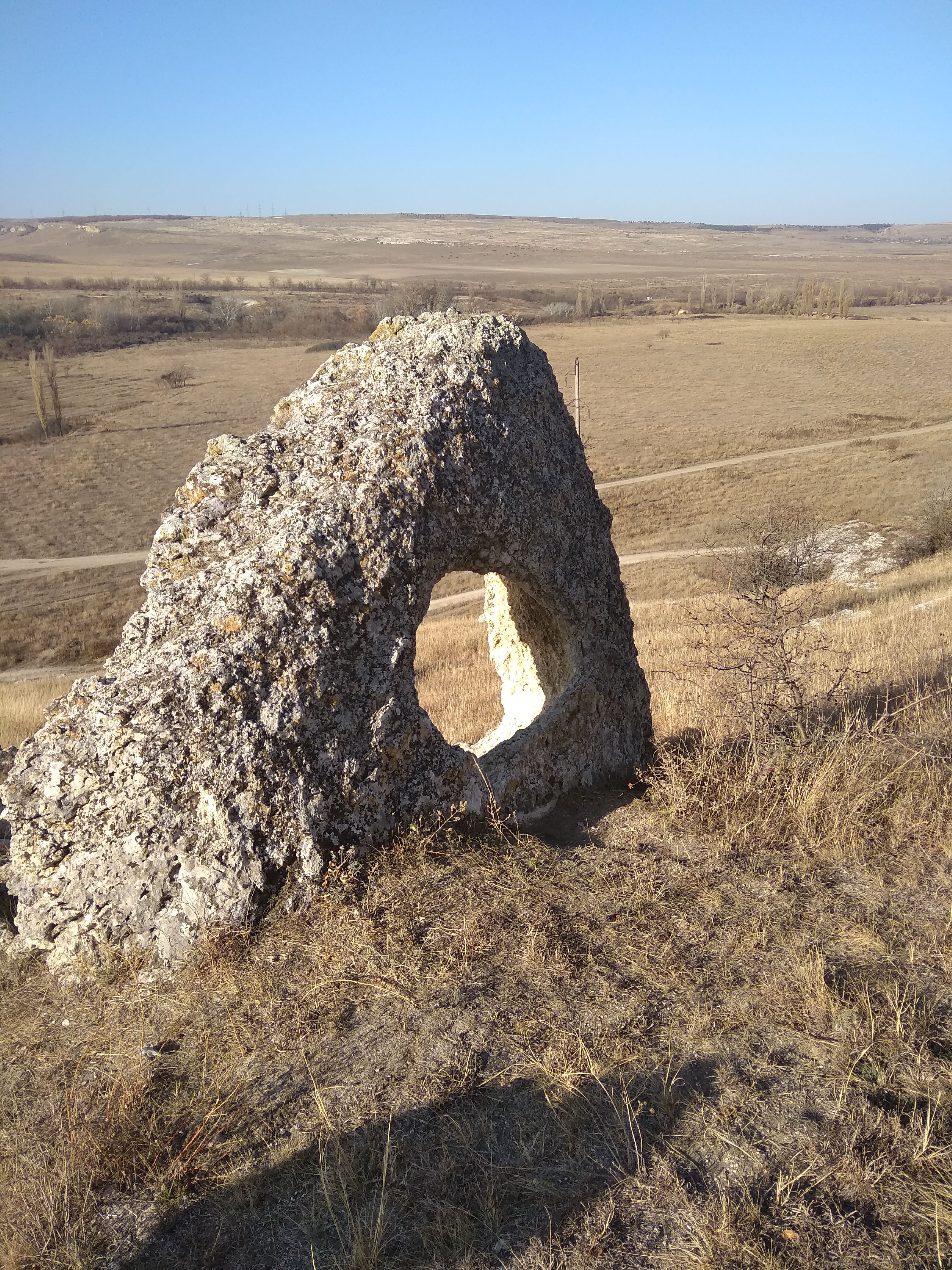 A unique rock formation with a hole stands in a vast natural field under clear skies.