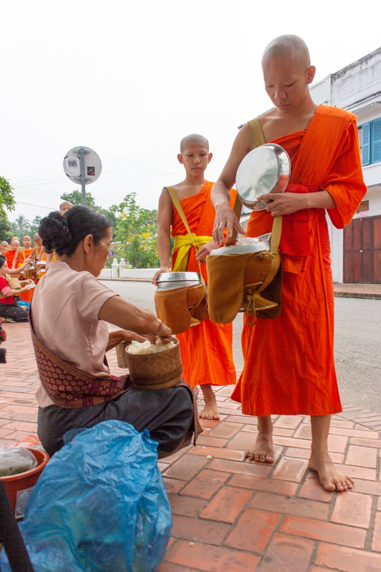 Monks Receiving Food Into Their Bags From A Woman On The Street