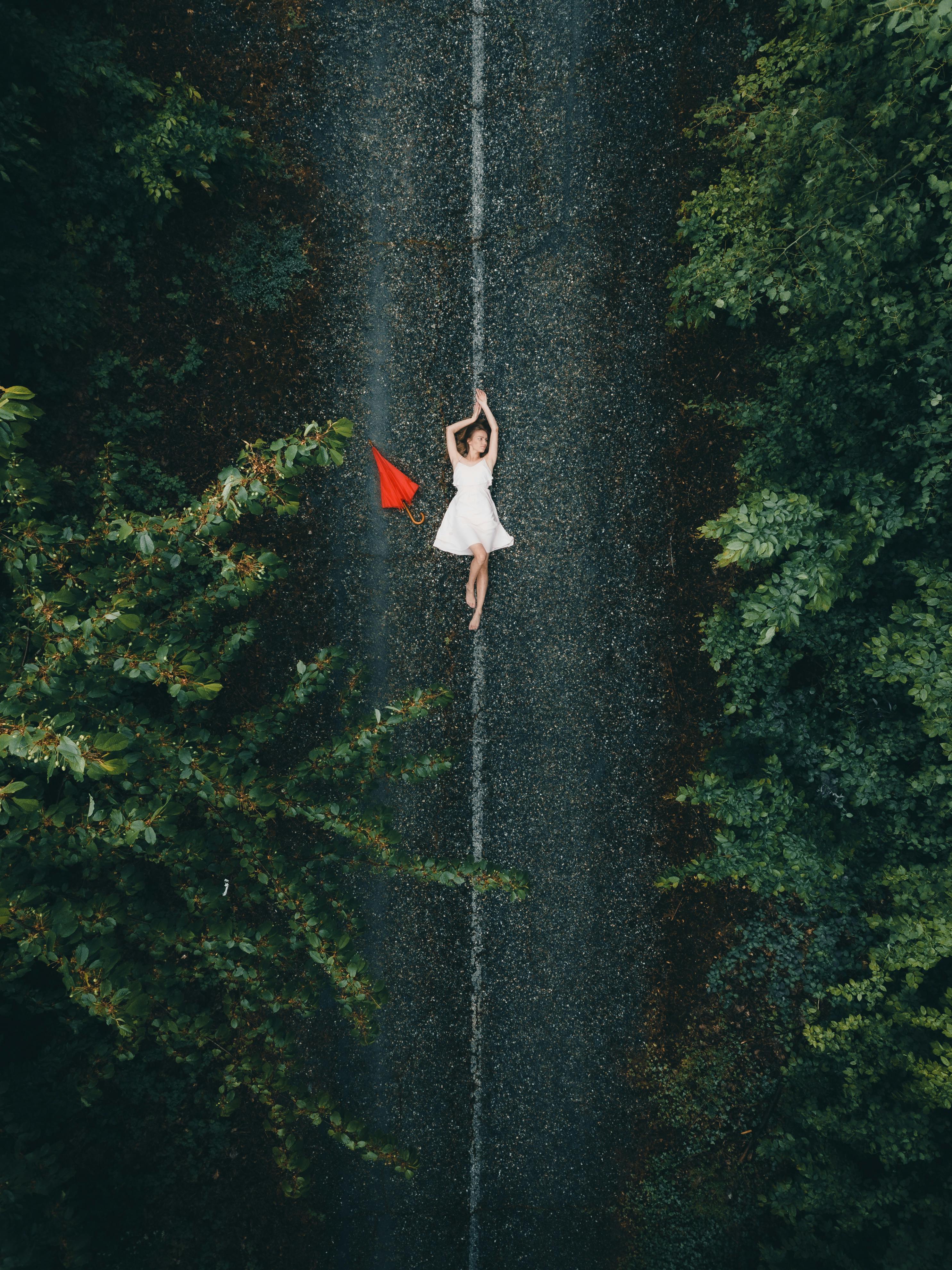 Aerial view of a woman in a white dress lying on a forest road with a red umbrella beside her.