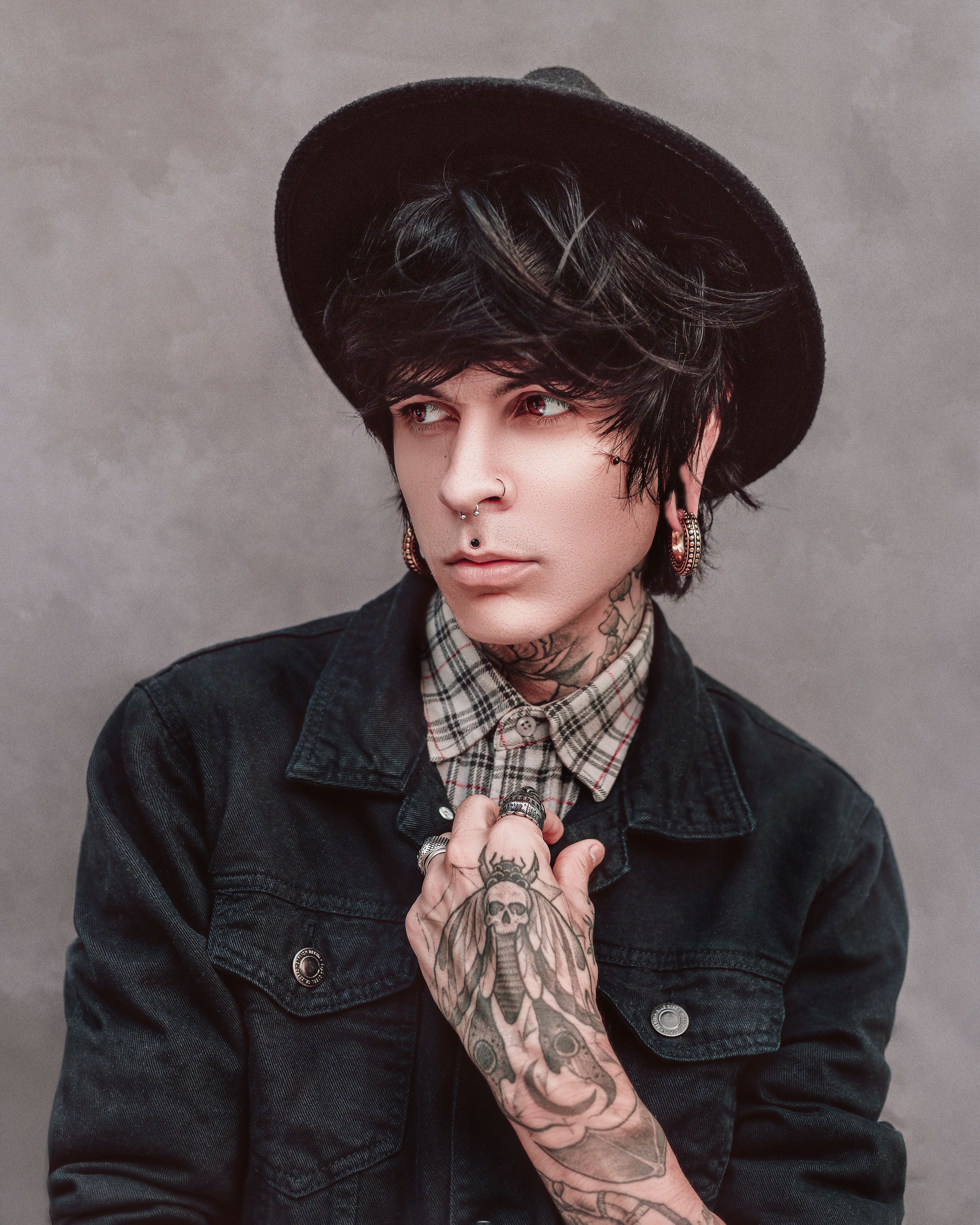 Tattooed person with piercings wearing a denim jacket and hat, showcasing unique style.