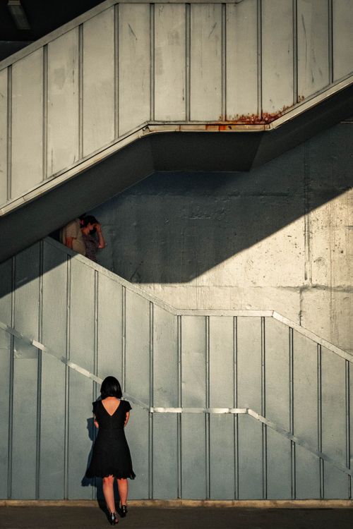 Free A woman in a black dress standing under an urban staircase, capturing city life essence. Stock Photo