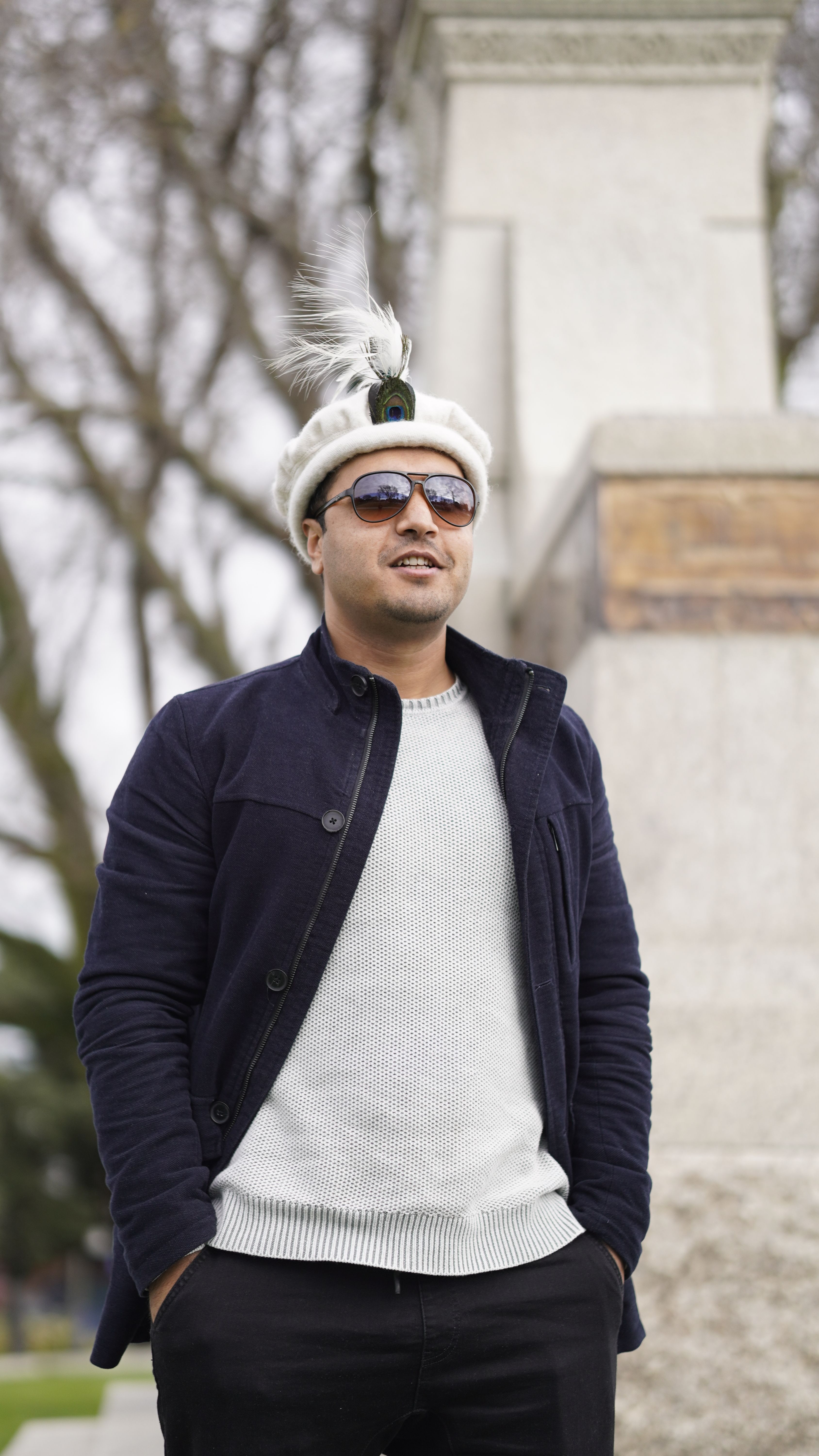 Man wearing feathered hat and sunglasses, standing outdoors against a stone structure.