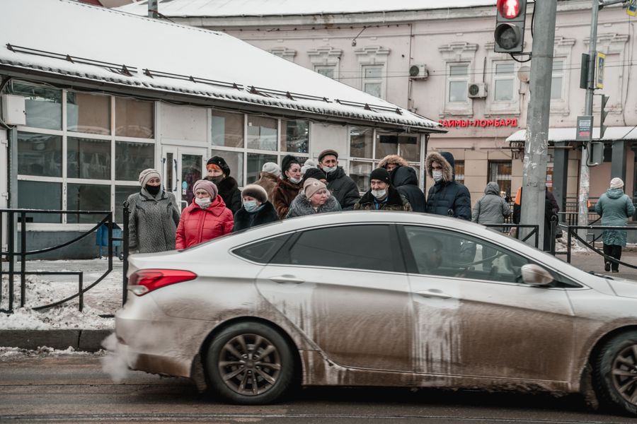 A winter city street with people wearing masks, jackets, and a passing car.