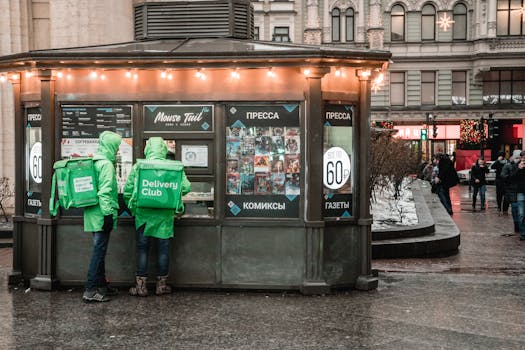Two delivery couriers in green jackets stand by a food stall at night.
