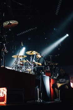 A drummer passionately plays a drum set on a dimly lit stage during a live concert.