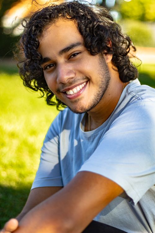 Free Portrait of a cheerful young man sitting on grass with sunlit background. Stock Photo