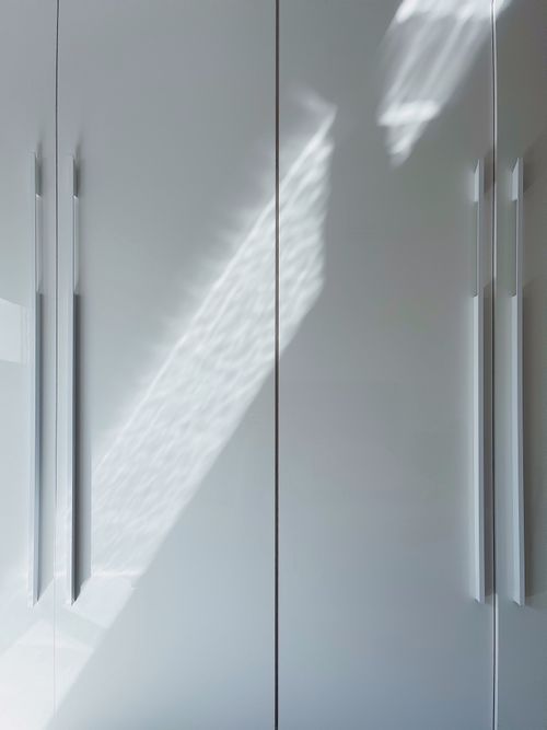 Free A modern minimalist white cabinet featuring sleek handles and striking sunlit shadows. Stock Photo