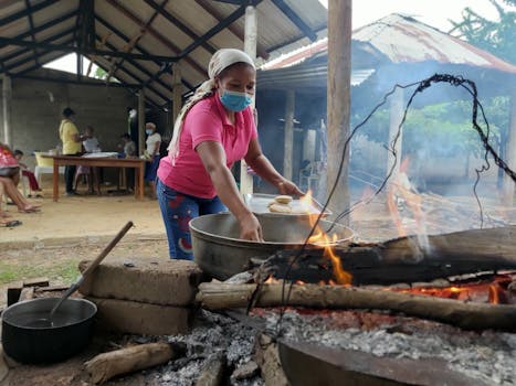 Woman in pink shirt cooking over open fire in rustic outdoor kitchen, wearing face mask.