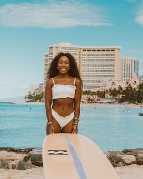Photo by Jess Loiterton Smiling woman in swimsuit on tropical beach with a surfboard, in front of a hotel, perfect for summer fun themes.