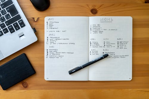 A Notebook with Pen on a Wooden Table Near the Laptop