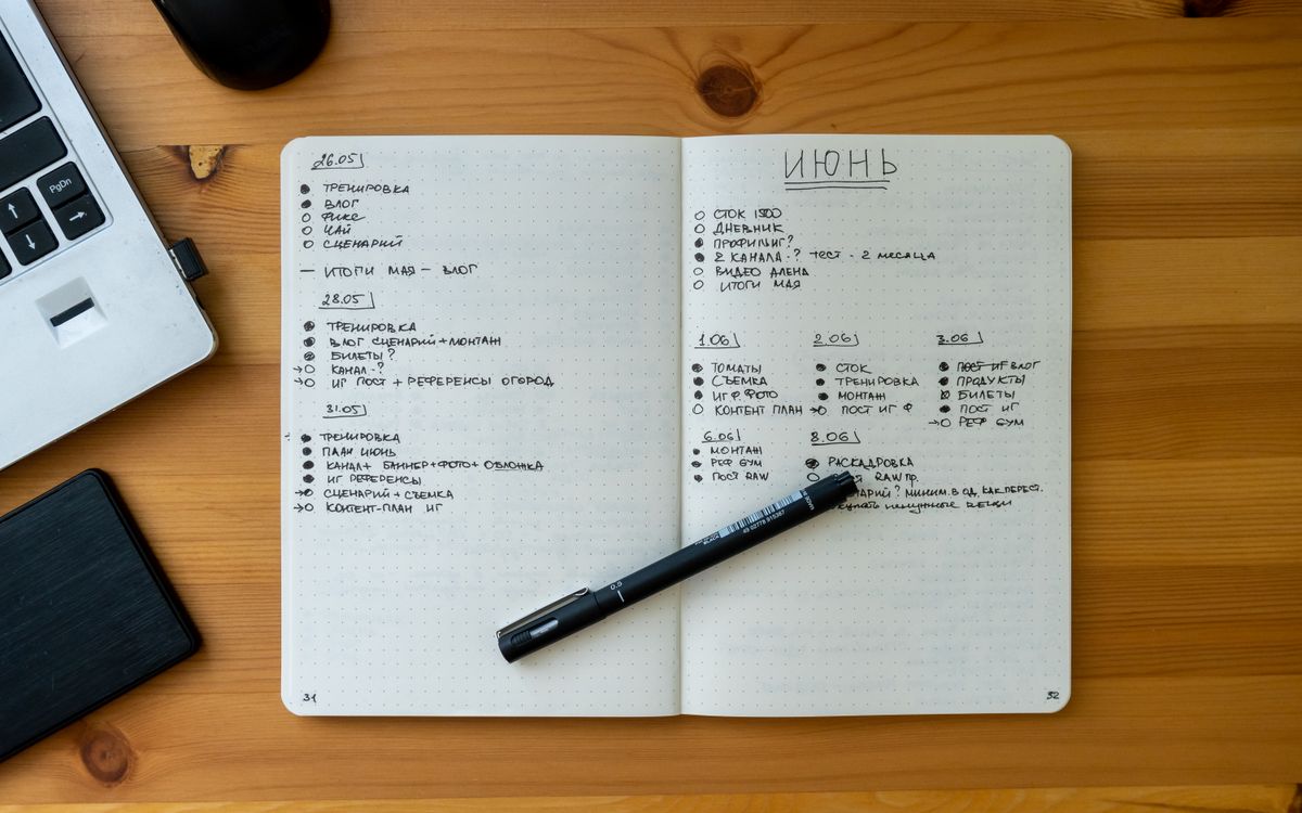 An open notebook with handwritten notes resting on a wooden desk beside a laptop and pen.