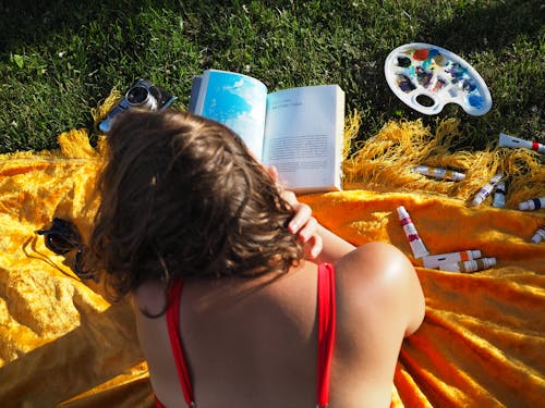 Free A Woman in Red Tank Reading a Book Near the Green Grass Stock Photo