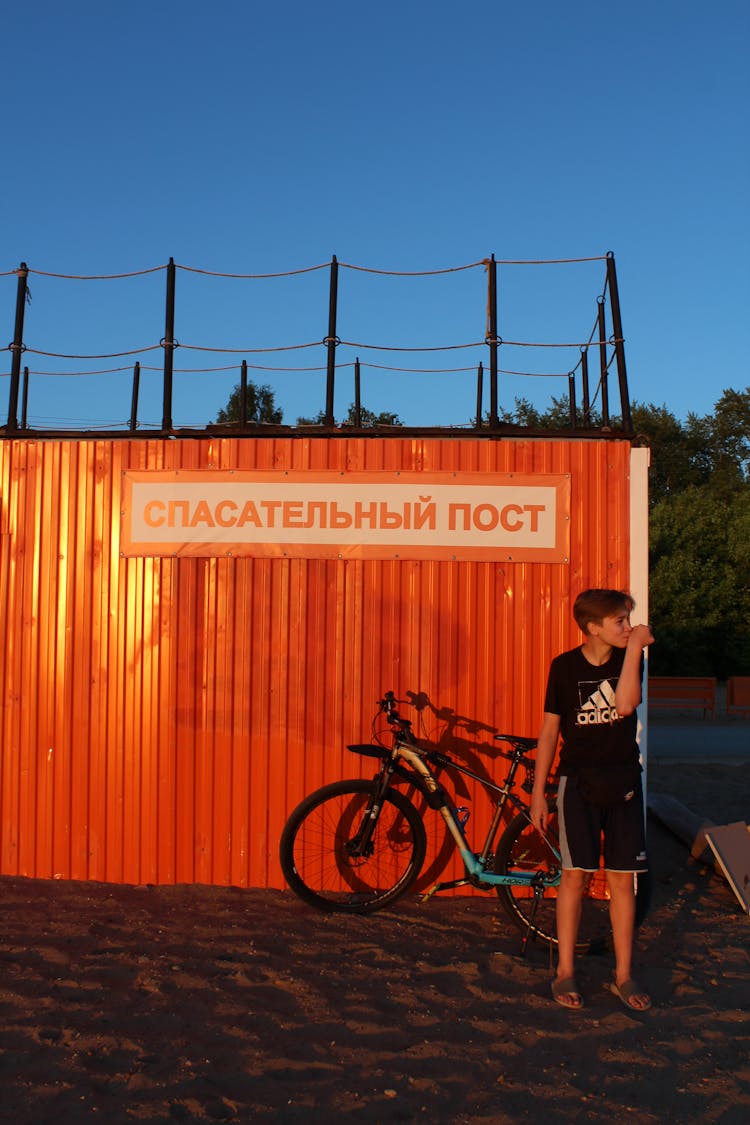 A Boy In Black Shirt Standing Near The Bicycle Parked On The Beach