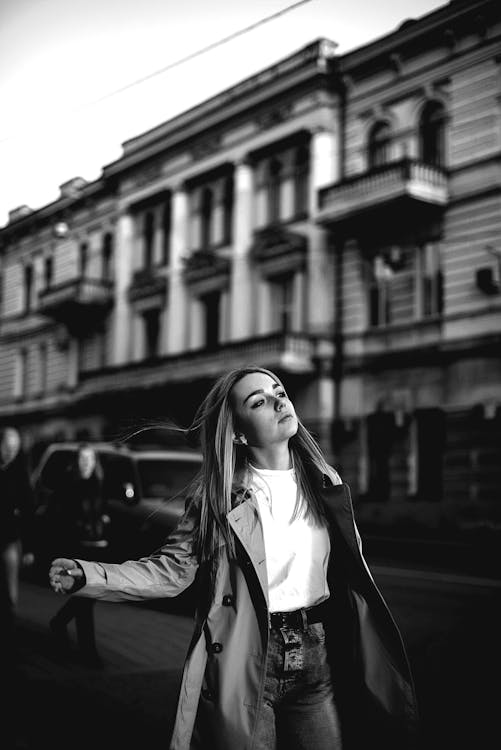 A Grayscale Photo of a Woman in Trench Coat Walking on the Street