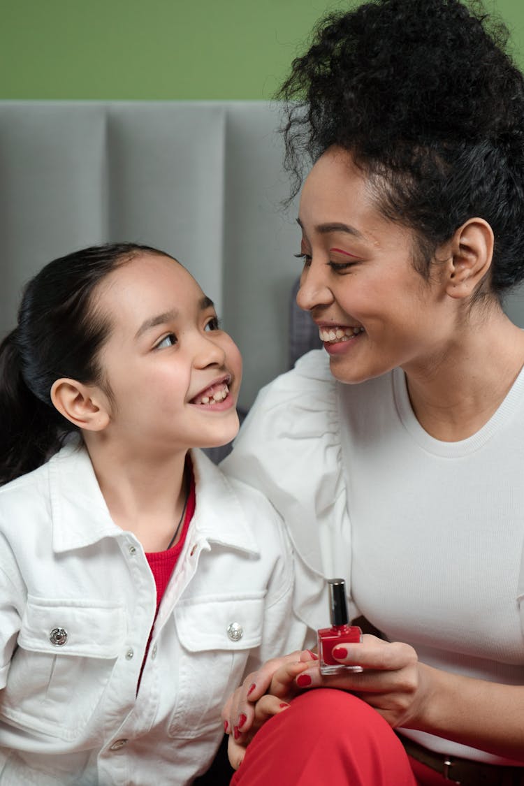 A Woman In White Shirt Holding A Nail Polish While Looking At Her Daughter