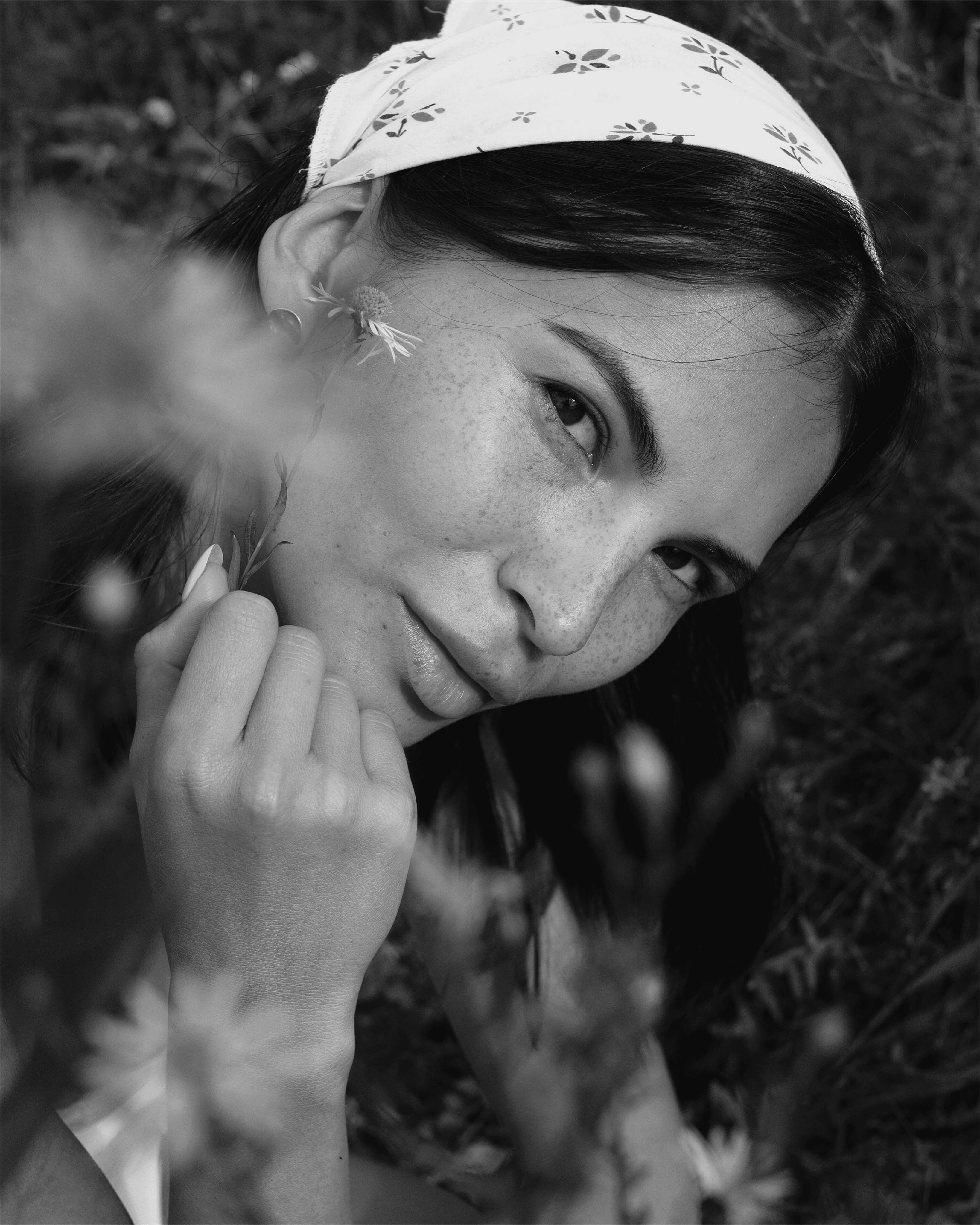 Artistic black and white portrait showing a woman with freckles and a headscarf amidst foliage.