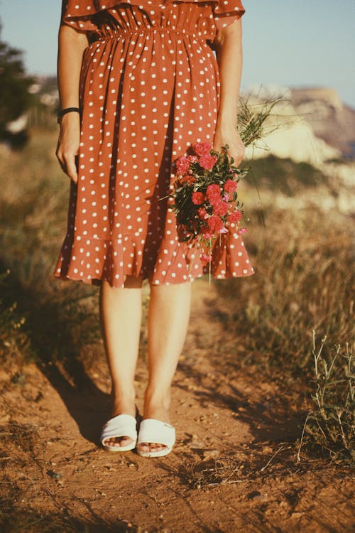 Person in Brown Polka Dot Dress Standing on the Ground While Holding Red Flowers