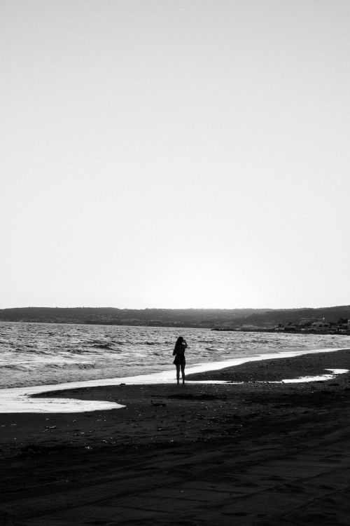 Free Black and white image of a solitary figure standing on a beach at dusk. Stock Photo