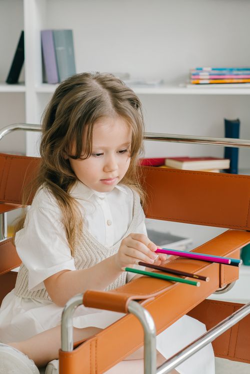 Free A young girl sits on a chair drawing with colored pencils, focused and serene. Stock Photo