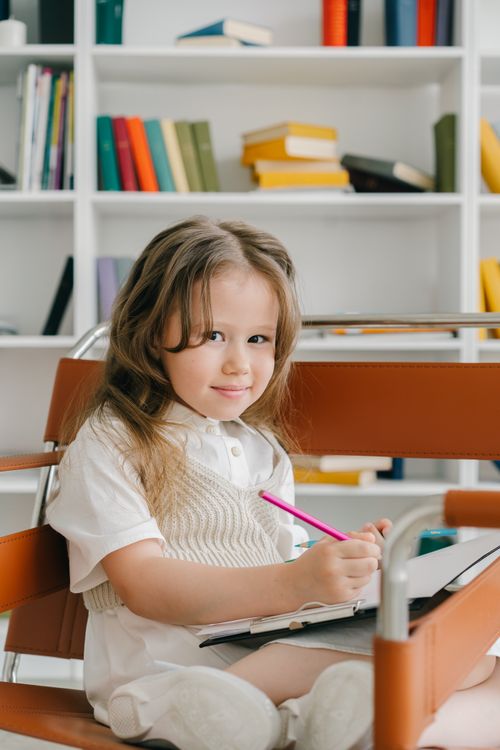 Free Cute young girl writing with a pink pencil in a cozy library setting. Stock Photo