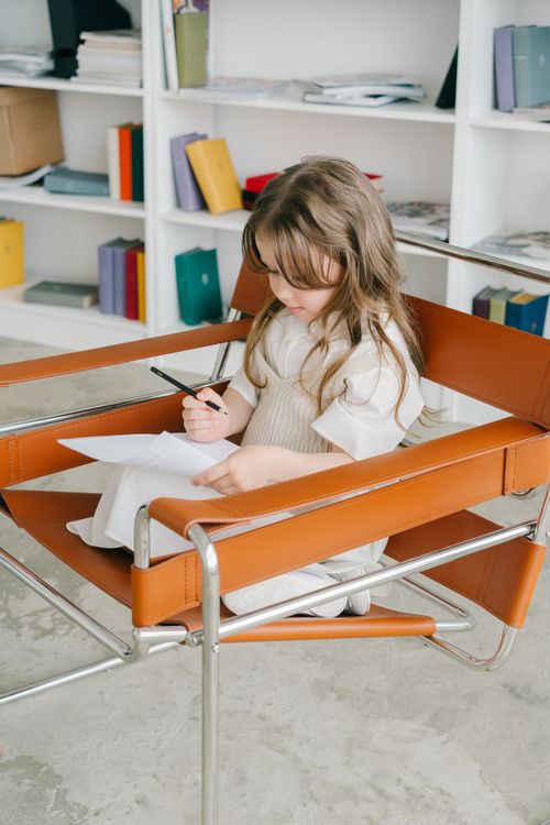 Free A young caucasian girl sits in a modern chair writing on paper, in a study with bookshelves. Stock Photo
