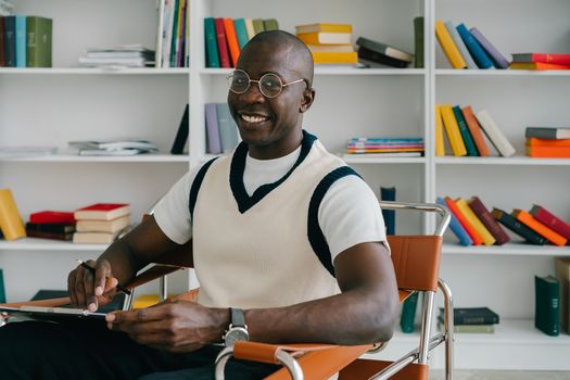 A cheerful man sitting in a library chair with a pen, surrounded by colorful books.