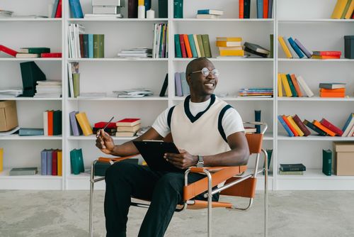 Free Man in a library setting sitting on a chair with a tablet and pen, smiling. Stock Photo