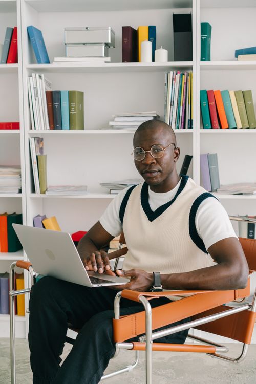 Free A man with eyeglasses working on a laptop in a modern office setting, seated by a bookshelf. Stock Photo