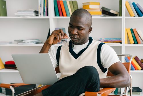 Free African American man sitting on chair using laptop in a library. Modern academic environment. Stock Photo