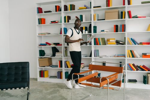 Free Stylish adult man standing with laptop in a modern library interior, surrounded by colorful bookshelves. Stock Photo