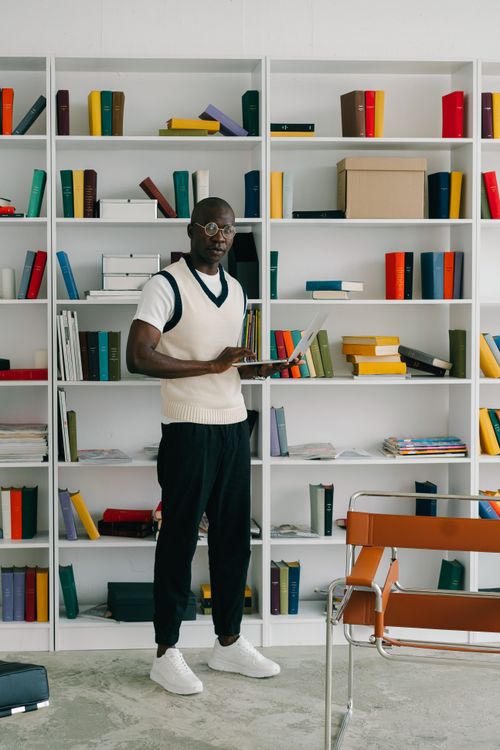 Free Man standing in a contemporary library setting, working with a laptop, surrounded by colorful books. Stock Photo