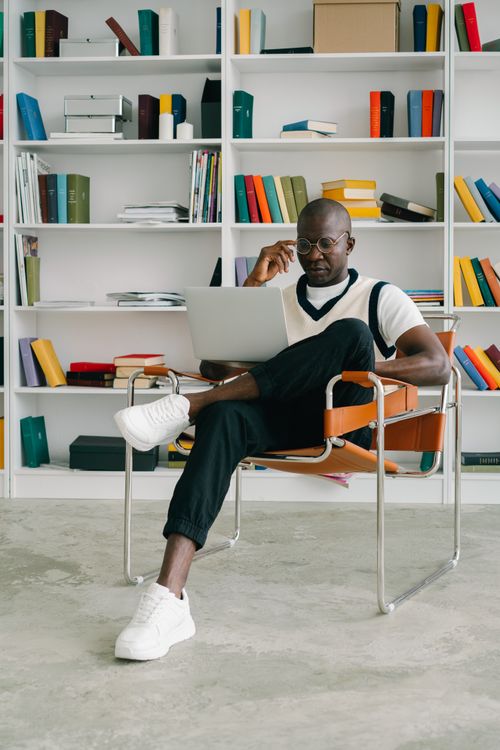 Free Stylish man in glasses working on laptop in a library setting, surrounded by colorful books on shelves. Stock Photo