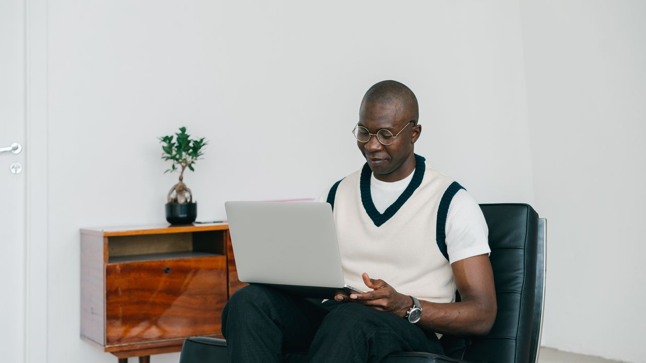 Black man in glasses focused on a laptop in a minimalist indoor space, seated on a black chair.