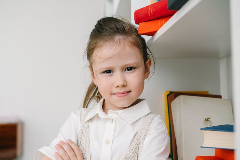 Cute young girl confidently posing with arms crossed next to a bookshelf. Cute young girl confidently posing with arms crossed next to a bookshelf.