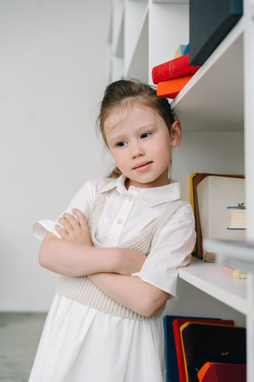 Free A young girl in a white dress leaning on a bookshelf, looking thoughtful. Stock Photo